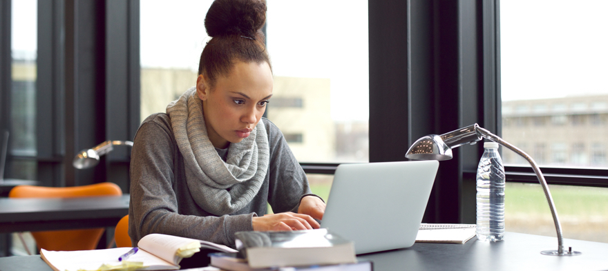 Young african american girl sitting in library searching information on laptop for her assignment. Young woman sitting at table surfing internet for notes for her study.